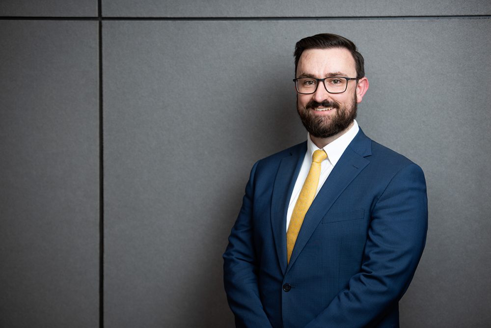 Blake Corporate type of portrait of a man standing. He is wearing a suit, white shirt and a tie.